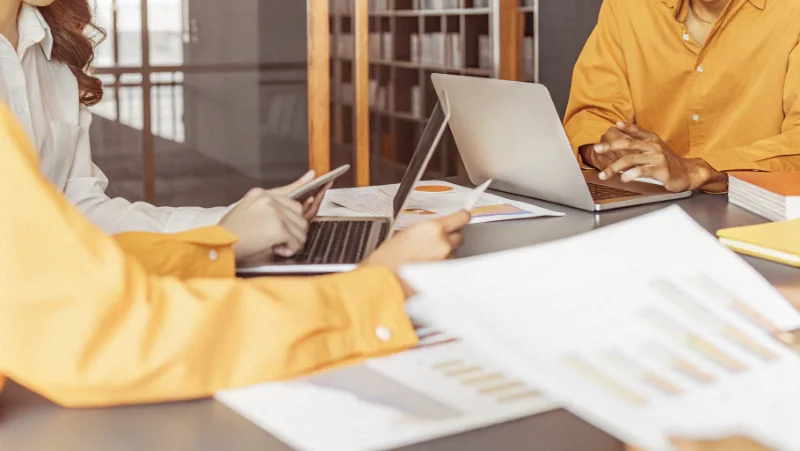 Team of researchers collaborating around table with laptops and data charts discussing quantitative analysis